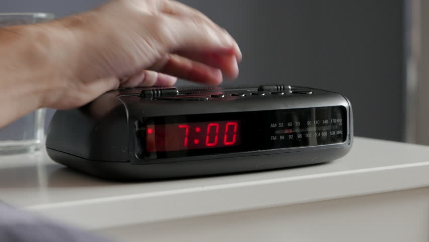 Alarm clock digital ringing 7am tired man pushes if of bed table 
